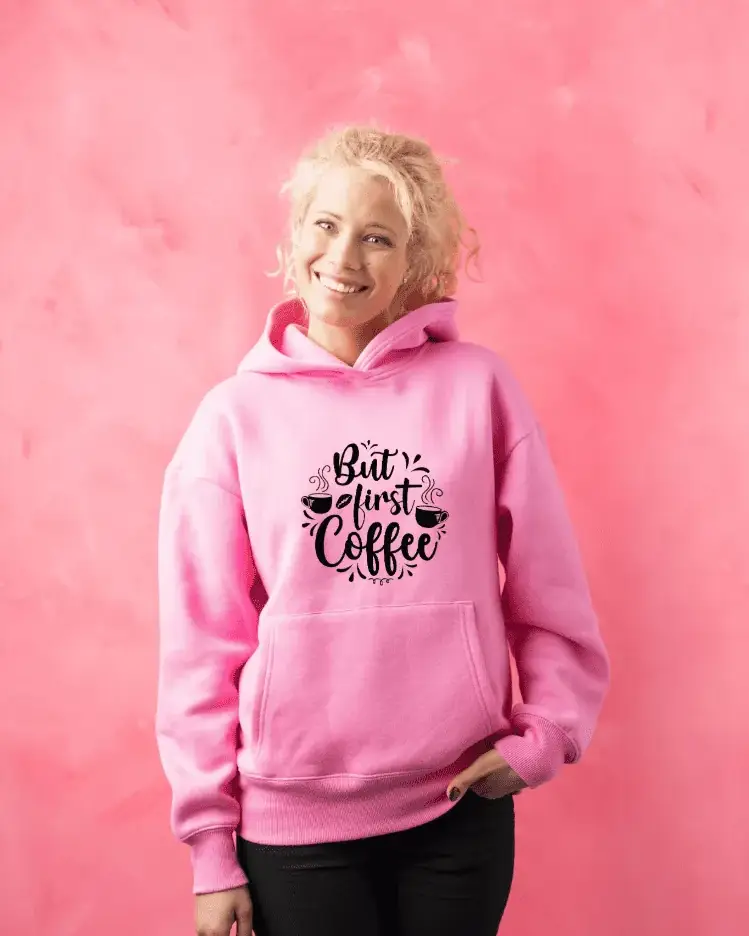 Blonde woman in pink hoodie with 'But first coffee' design Smiling woman in a pink hoodie with 'But first coffee' text and coffee motif, standing in front of a pink wall – customize your hoodie