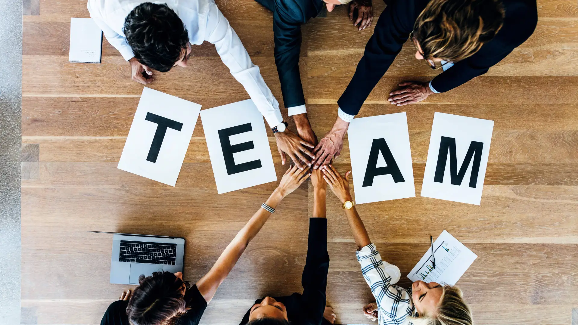 Teamwork with Company T-Shirts – Team Building through Uniform Clothing Teamwork with corporate clothing – hands stacked above the word 'Team', T-shirts for team building.