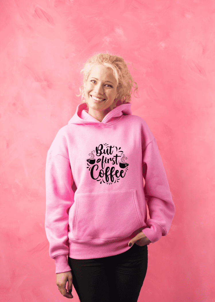 Lachende Frau trägt pinken Hoodie mit 'But first coffee'-Spruch und Kaffeemotiv, steht vor pinker Wand – Hoodie individuell gestalten