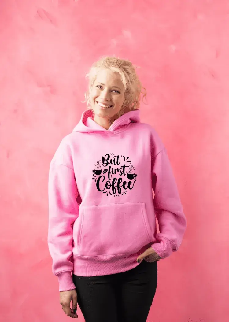 Lachende Frau trägt pinken Hoodie mit 'But first coffee'-Spruch und Kaffeemotiv, steht vor pinker Wand – Hoodie individuell gestalten
