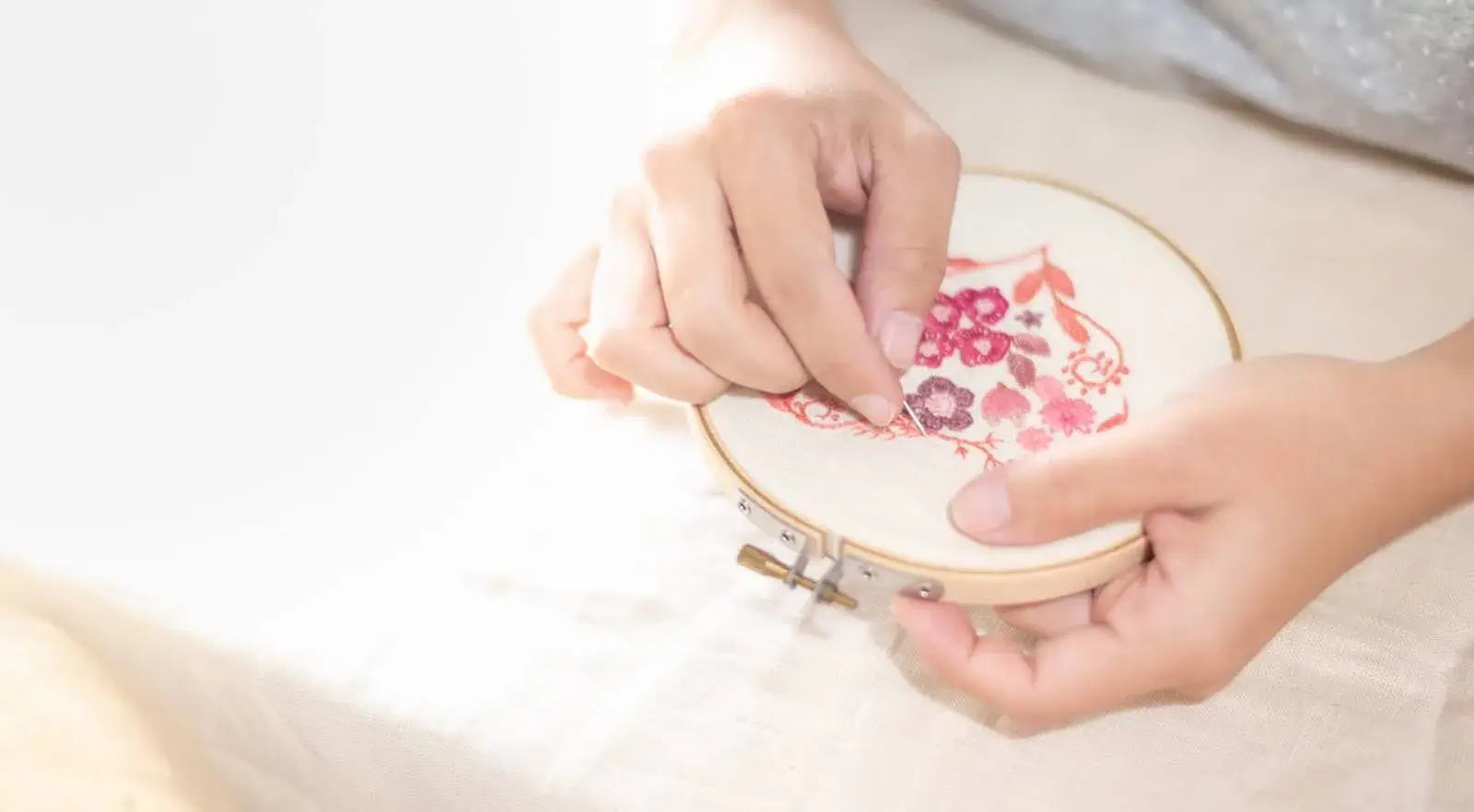 Frau stickt ein rosa Blumenmuster mit Nadel und Stickrahmen auf Stoff – filigrane Handarbeit in der Entstehung